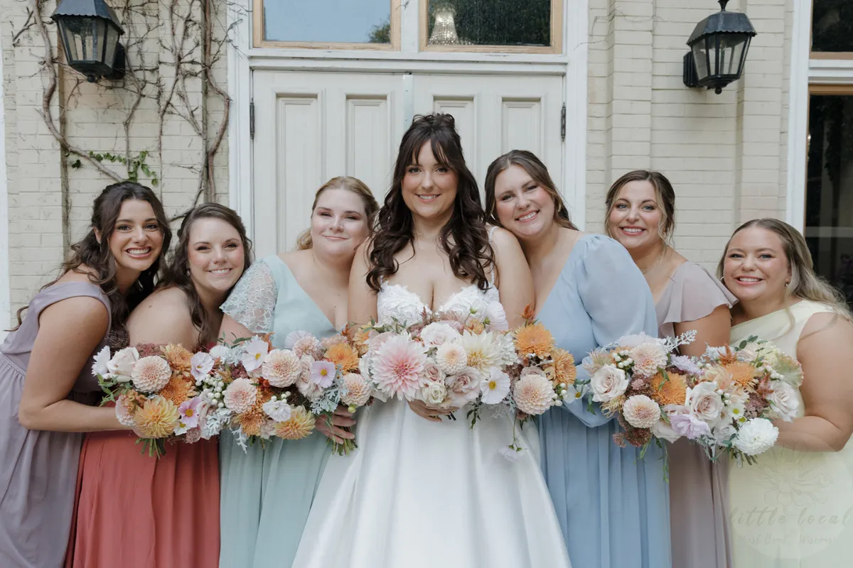 Bride and bridesmaids holding pastel bouquets against cream city brick wall with ivy
